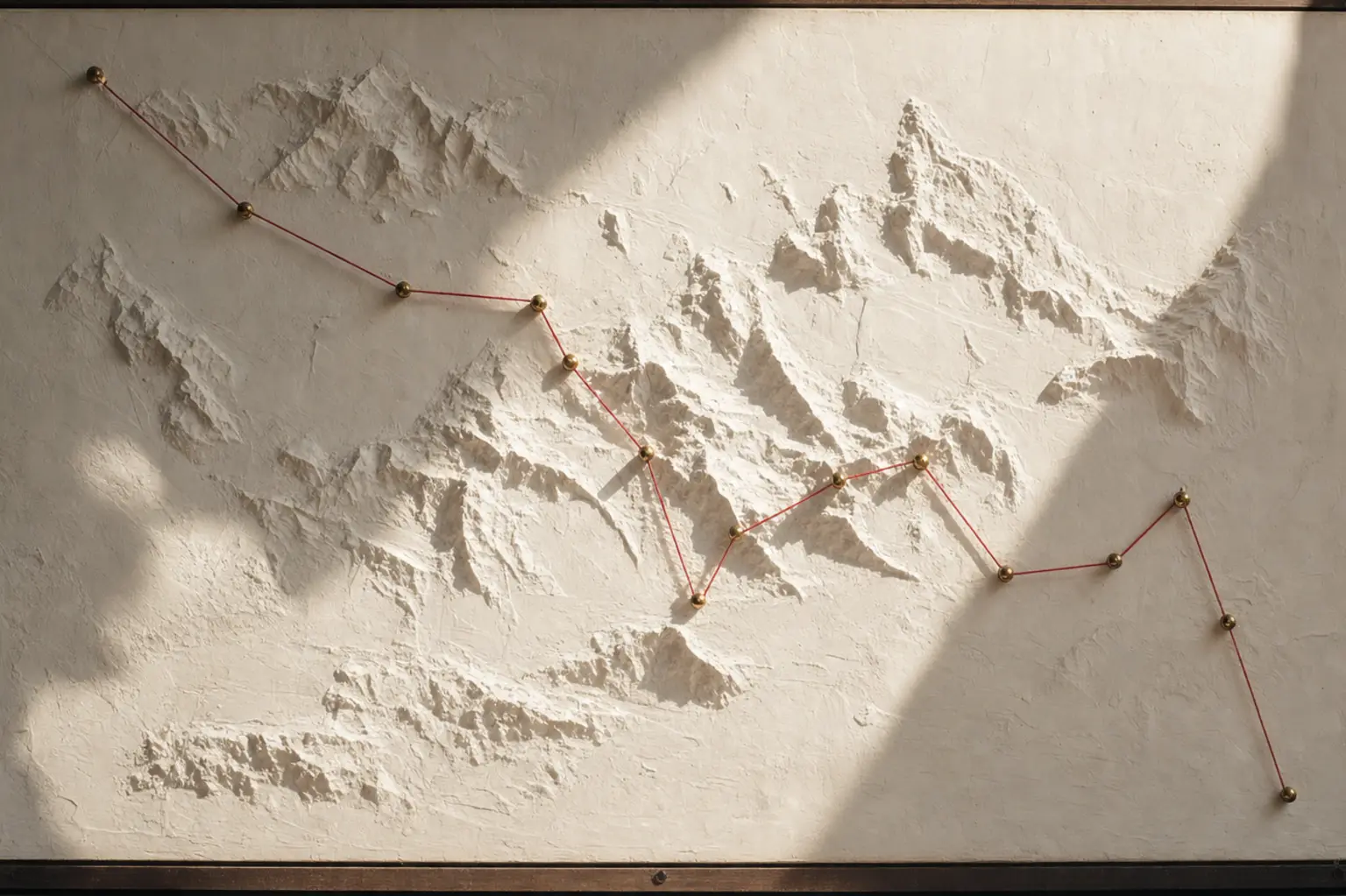 A red thread pulled taut between brass pins across a plaster-white relief map under hard noon light.